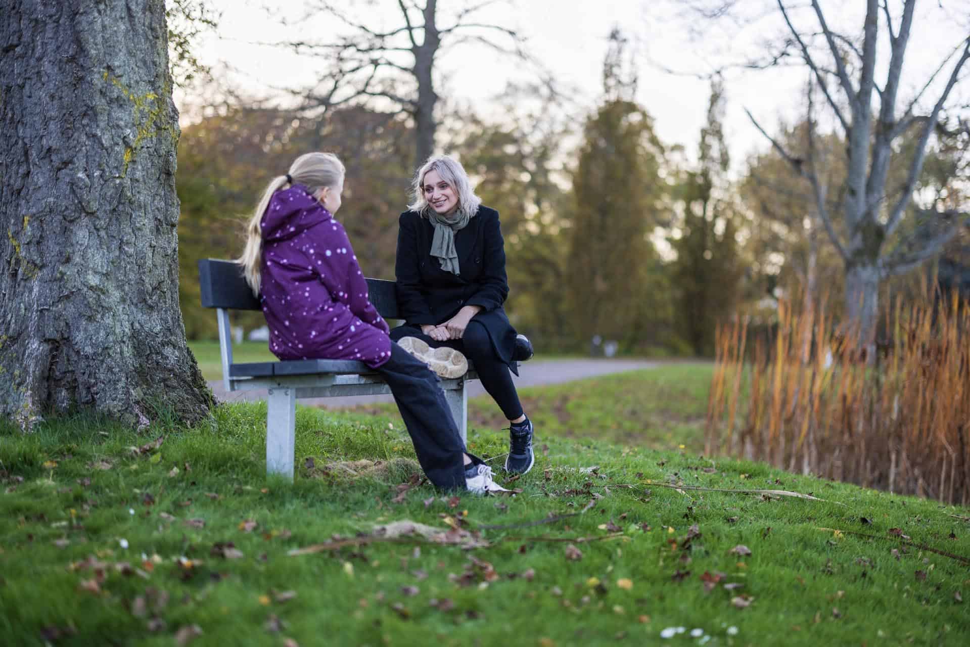 Kvinde og et barn sidder overfor hinanden i samtale ved en bænk på en græsplæne.