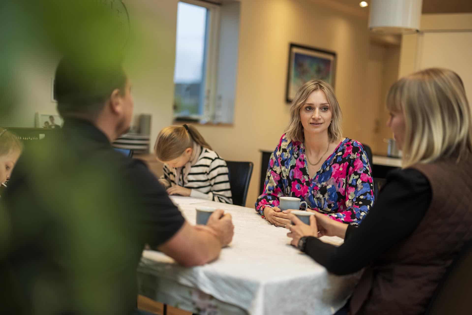 Fem personer sidder omkring et spisebord dækket med en hvid dug og taler sammen.